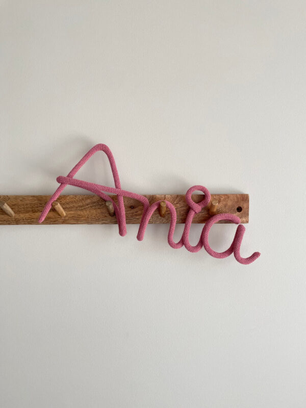 Napis ze sznurka Ania wisi na ścianie na drewnianym wieszaku.