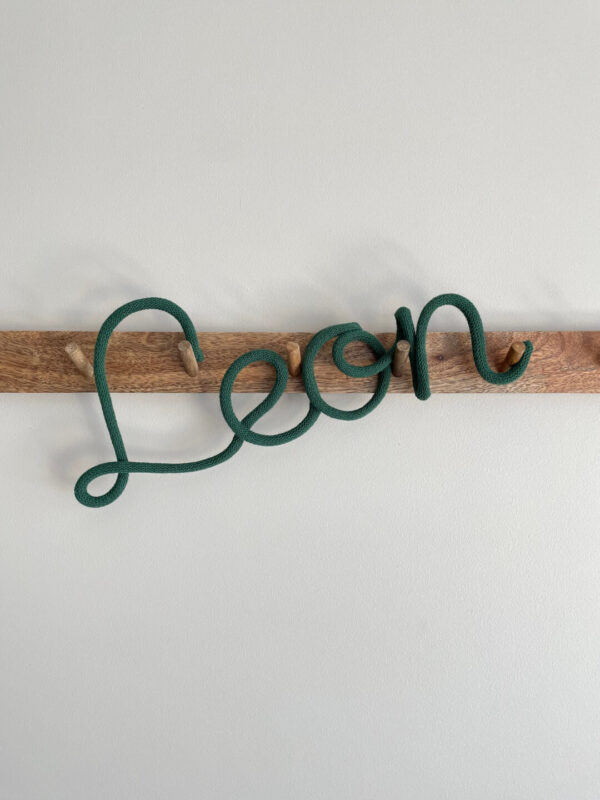Napis ze sznurka Leon wisi na ścianie na wieszaku z drewna.