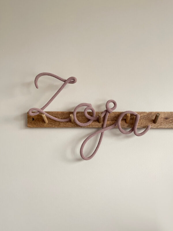Napis ze sznurka Zoja wisi na drewnianym wieszaku ściennym.