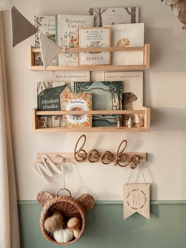 Zdjęcie przedstawia ścianę w pokoju dziecięcym, na której wiszą drewniane półki, na których stoją książki dla dzieci, a poniżej wisi napis ze sznurka books.