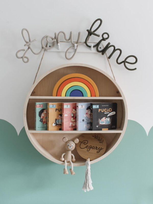 Napis storytime wisi na ścianie nad drewnianą okrągłą półeczką w pokoju dziecięcym. Na półeczce stoją książki dla dzieci, drewniana tęcza i inne dekoracje dziecięce.