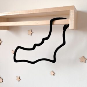 Dekoracja dziecięca ze sznurka w kształcie nietoperza wisi na drewnianym wieszaku ściennym. W tle na ścianie zamocowane są małe drewniane gwiazdki.