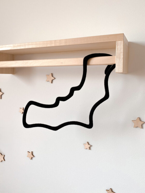 Dekoracja dziecięca ze sznurka w kształcie nietoperza wisi na drewnianym wieszaku ściennym. W tle na ścianie zamocowane są małe drewniane gwiazdki.