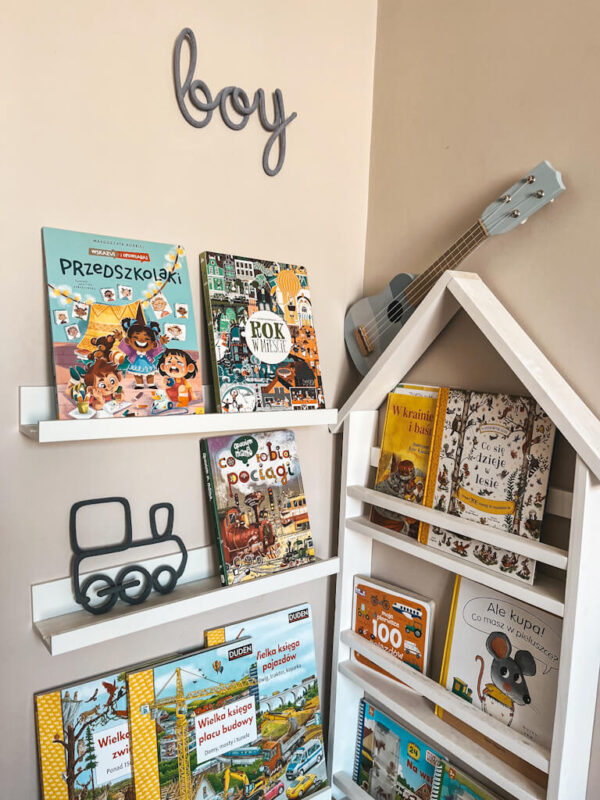 Dekoracja ze sznurka w kształcie pociągu stoi na półce z książkami w pokoju dziecka. Napis ze sznurka boy wisi na ścianie nad książkami dla dzieci.