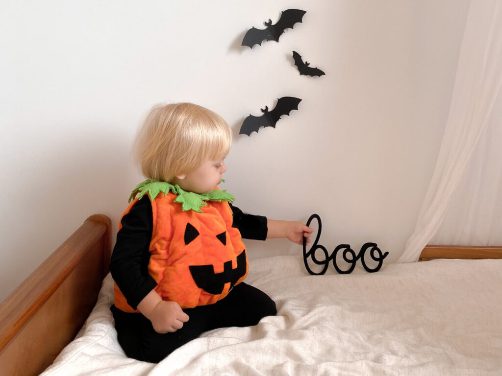 Małe dziecko ubrane w przebranie dyni na Halloween siedzi na łóżku i trzyma w dłoni czarny napis ze sznurka boo.