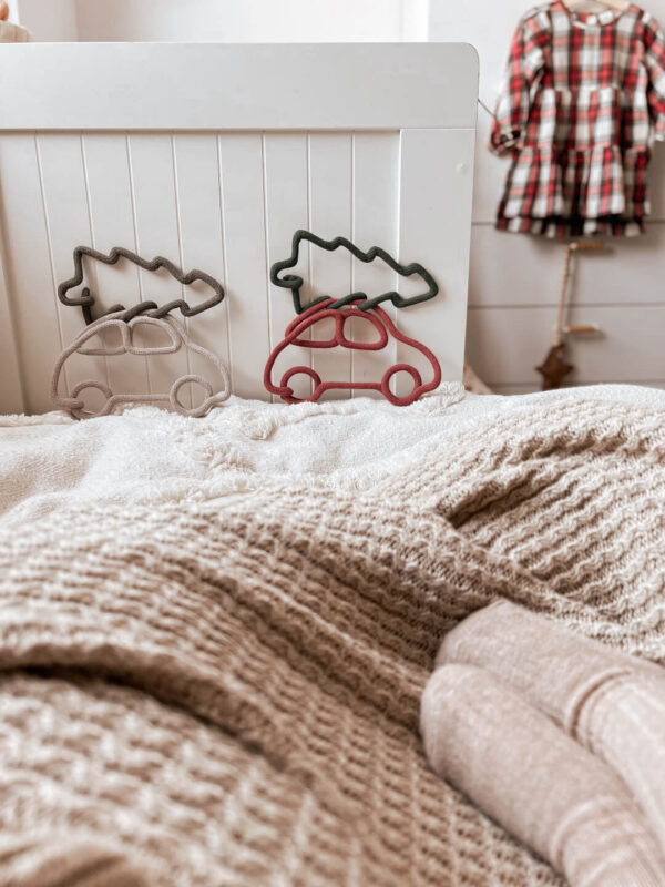 Świąteczne ozdoby ze sznurka w kształcie autek z choinkami na dachu stoją oparte o ramę łóżka w pokoju dziecka.