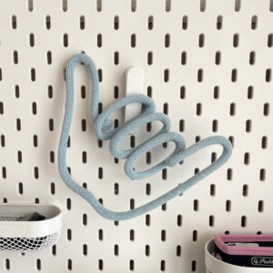 Dekoracja ze sznurka w kształcie gestu shaka wisi na tablicy ściennej IKEA SKADIS,